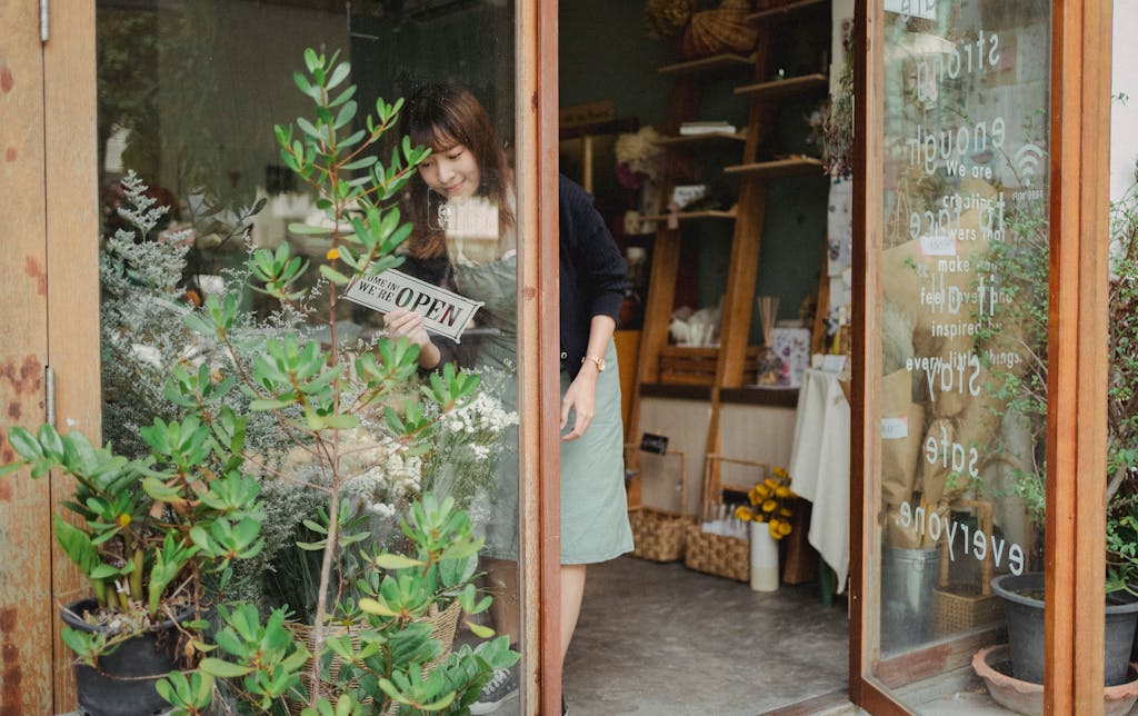 Positive young Asian female florist in apron standing near entrance and turning signboard on glass wall while working in modern floristry shop